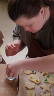 Down sendromlu bir kızın dikey yüksek açı çekimi ve kadın bakıcı fırın kağıdındaki kurabiyelere renkli şeker damlaları ekliyor.