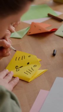 Down sendromlu bir kızın masa başında öğretmeninin yanında oturmuş kağıt işçiliği yaparken origami hayvanının yüzünü çizdiği dikey yakın çekim.