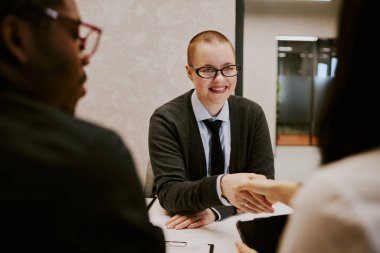 Buluşma sırasında iş ortağıyla el sıkışırken neşeli, kısa saçlı genç bir kadının omuz üstü fotoğrafı.