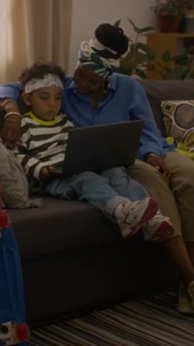 Afro-Amerikan kökenli bir kadın ve küçük oğlunun evdeki kanepede otururken dizüstü bilgisayarda video izlediği dikey bir fotoğraf.