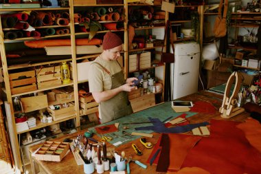 High angle shot of male tailor holding deep blue colored leather workpiece and looking at it at work in fully equipped atelier clipart