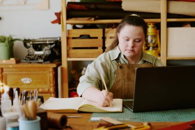 Female tailor with down syndrome taking notes while sitting at old shabby table with sewing tools on top clipart