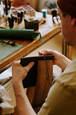 Over shoulder shot of man sitting at table and sewing stitches on leather clipart