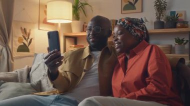 Afrikalı Amerikalı genç bir çift arkadaşlarıyla video görüşmesi yapıyor. Oturma odasında kanepede otururken gülümsüyor ve aktif bir şekilde konuşuyorlar.