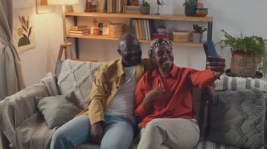 Afro-Amerikalı çift oturma odasında kanepede otururken selfie çekiyorlar, sonra da telefondaki fotoğraflara bakıyorlar.