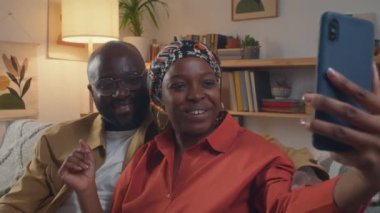 Genç siyahi kadın oturma odasında kanepede otururken kocasıyla selfie çekiyor, sonra gülümsüyor ve akıllı telefondan fotoğraf tartışıyor.