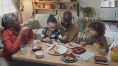 Afro-Amerikalı üvey aileler ve iki beyaz çocuk yemek masasında aile kahvaltısı yaparken birbirleriyle konuşuyorlar.