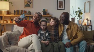 Genç Afrikalı Amerikalı anne, oturma odasındaki kanepede otururken iki beyaz çocuk ve siyah kocasıyla birlikte akıllı telefondan selfie çekiyor.