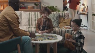 Siyahi bir adam, evlatlık beyaz oğullarla masa oyunu oynuyor, sonra karısı bu ziyaretçiyle oturma odasına girerken ve dairesini gösterirken kadın sosyal hizmet görevlisiyle tokalaşıyor.