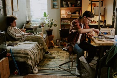 Young college student sitting at work desk and repairing laptop while his friend sitting on bed and playing computer games clipart