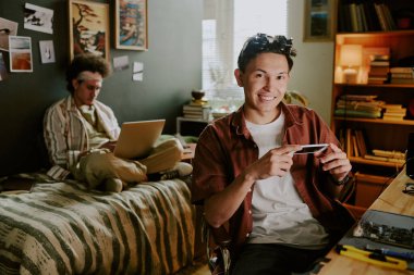 Smiling university student looking directly at camera while repairing his phone, his friend sitting on bed and coding clipart