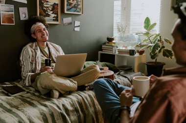 Curly-haired student sitting with laptop on bed and loathing out loud while his roomie telling joke and drinking coffee clipart