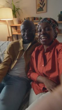 Afro-Amerikalı genç eşlerin el sallayıp kameraya bakarken arkadaşlarının evdeki kanepede otururken görüntülerini dikey çekiyoruz.