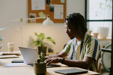 Genç Afrikalı Amerikalı bir adam modern çevre dostu ofiste otururken dizüstü bilgisayarda daktilo kullanıyor.