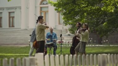 Klasik müzik enstrümanları çalan ve parktaki yeşil çimlerde açık havada eğlenen neşeli genç müzisyenlerden oluşan bir grup.