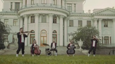 Çello, davul ve keman çalan profesyonel müzisyenlerden oluşan geniş bir sahne. Tarihi binanın önündeki yeşil çimlerde klasik müzik konseri veriyorlar.