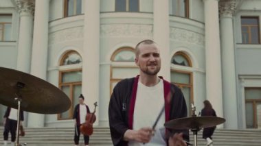 Dışarda davul çalan etkileyici erkek müzisyenin çekimlerinden uzaklaşırken grup arkadaşları klasik müzik enstrümanlarıyla tarihi binanın merdivenlerinde dans ediyorlar.