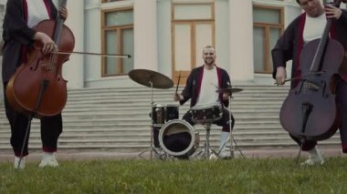 Neşeli erkek davulcu, parkta açık hava müzik performansı yaparken iki çellocu eşliğinde davul çalıyor.