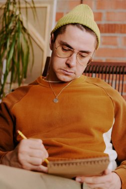 Eskiz defterine çizim yapmaya odaklanmış gözlüklü genç beyaz erkek ressamın dikey orta pozu.