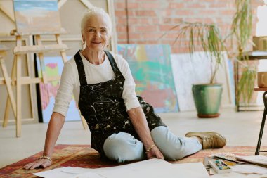 Modern sanat stüdyosunda önlük giymiş, yerde oturmuş kameraya gülümseyen yaratıcı beyaz kadın portresi.