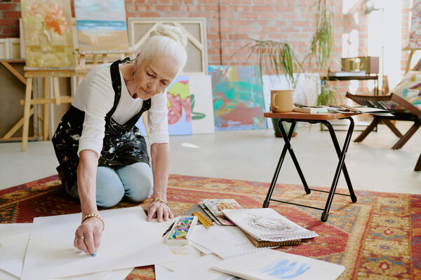 Senior Caucasian female artist sitting on floor in her studio and drawing with soft pastels, copy space