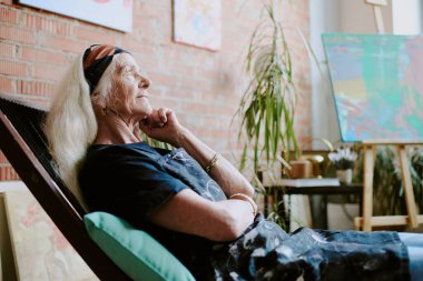 Stüdyoda resim yaptıktan sonra tahta sandalyede dinlenen beyaz kadın ressamın yan görüntüsü.