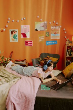 Engelli genç kızın dikey yüksek açılı görüntüsü şık yatak odasında dinleniyor, müzik dinliyor ve dizüstü bilgisayardaki web sitelerini karıştırıyor.