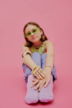 Protez kollu, şık elbiseli pembe arka planda oturan kendine güvenen bir genç kızın dikey stüdyo çekimi.