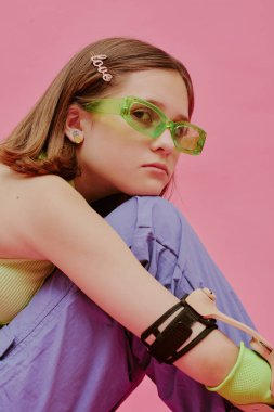 Kafkasyalı engelli genç kızın dikey yakın çekimi. Gözlük takıyor, pembe arka planda oturuyor.