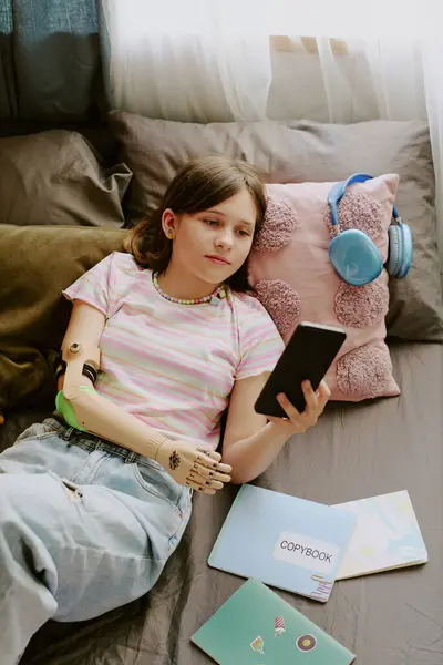 stock image Vertical from above view shot of modern teen girl with disability reading book on her smartphone while relaxing on bed