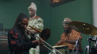 Afrikalı Amerikalı bir adam trombon çalarken erkek orkestra arkadaşı konga ve kadın vokalist üzerinde Guiro ile prova yapıyor.