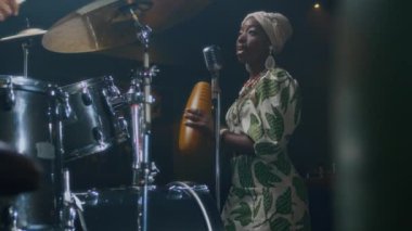 Barda tanınmayan bir davulcuyla prova yaparken şarkı söyleyen ve guiro çalan siyahi genç bir kadın.