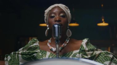 Barda mikrofonun önünde durup kameraya bakarak şarkı söyleyen Afrikalı Amerikalı kadın vokalist.