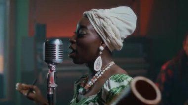 Afrikalı Amerikalı kadın vokalist mikrofonun önünde şarkı söylüyor. Müzik grubuyla birlikte barda, arka planda erkek tromboncu.