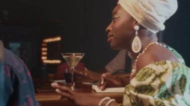 Afro-Amerikalı bir kadın barda oturmuş içki ve not defterleriyle birlikte yeni şarkılar bestelerken heyecanlı bir şekilde grup arkadaşlarıyla konuşuyor.