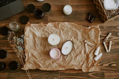 A rustic setup showcasing homemade candle-making process with different essential oils and dried flowers on brown paper, jars, and tools arranged on wooden surface clipart