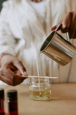 İnsanlar dikkatlice eritilmiş balmumunu metal bir sürahiden mum kavanozuna dökerek arka planda kırmızı şişelerle mum yapımı sürecine göz atarlar.