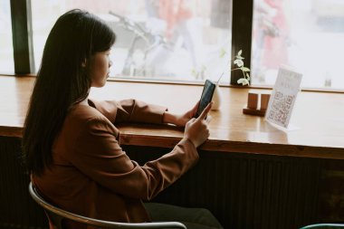 Asian woman sitting by window using smartphone and looking out at street view, creating ambiance of contemplation and productivity in quiet cafe setting clipart