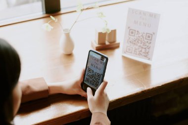 Konforlu kahve dükkanındaki akıllı telefondaki soru cevap kodunu tarayan kişi. Pencerelerden doğal aydınlatma ve ahşap masadaki küçük bitki.