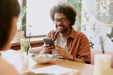 Restoran masasında oturan gülümseyen genç adam akıllı telefonuyla sohbet ederken yemek ve içeceğin tadını çıkarıyor.