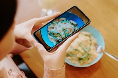 Lezzetli kremalı makarna tabağının fotoğrafını çeken kişi akıllı telefonuyla taze otlarla süslenmiş. Başının arkası görünürdü, yemek yakalamaya odaklanmıştık.
