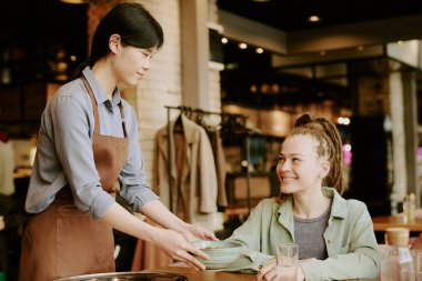 Asyalı garson, Kafe 'de sıcak bir ortam ve personel ile müşteri arasında dostça bir atmosfer ve olumlu bir etkileşim yaratan, rahat bir ortamda genç beyaz bir kadına hizmet veriyor.