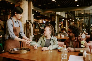 Garson sıcak bir ortam yaratmak için rahat restorantta gülümseyen çifte yemek getiriyor. Modern ve davetkar iç mekanlarla çevrili çeşitli gruplar yemeğin tadını çıkarıyor.