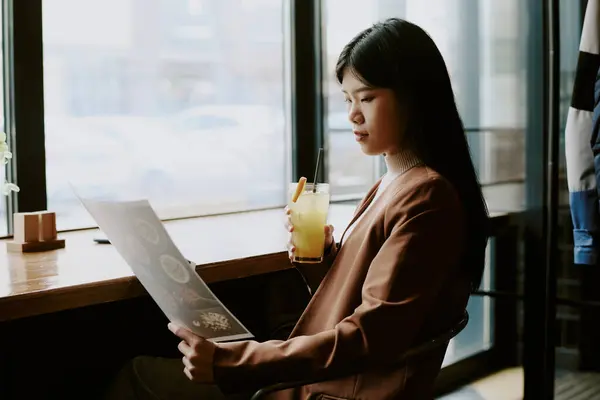 stock image Woman in brown blazer sitting by window, holding glass of lemonade and reading menu, in intimate cafe with wooden table and natural lighting