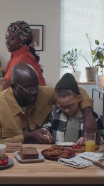 Afrika kökenli Amerikalı babanın evlat edinilmiş beyaz evladının domatesli sandviç yapmasına ve ekmeğe peynir sürmesine yardım ederken çekilmiş dikey bir fotoğrafı. Anne yemek masasında portakal suyuyla geçerken görülüyor.