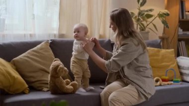 Orta boy beyaz bir kadın, annesinin desteğiyle kanepede duran sevimli bir kızın elini tutuyor ve ilk adımlarına hazırlanıyor.