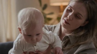 Beyaz tenli genç bir annenin orta boy yakın çekimi. Sarışın, sevimli bir bebeği dikkatlice kucağına alıyor. Arka planda rahat bir ortam var.