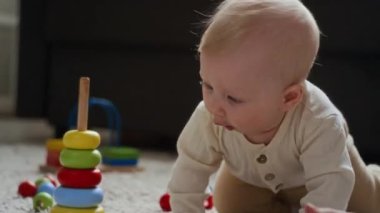 Meraklı, beyaz bir bebeğin halı zemininde renkli oyuncaklarla oynaması, yeni nesneleri keşfederken motor becerilerini geliştirmesi. Ekran dışındaki kadının çocukla etkileşimi