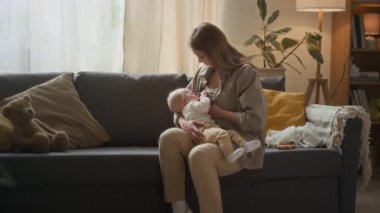 Evdeki rahat gri koltukta oturan beyaz bir kadının bebeğini büyümesi ve gelişmesi için emzirmesi.