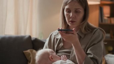 Orta boy beyaz anne elinde akıllı telefon ile sallanırken ve oğlunu emzirirken sesli mesaj kaydediyor.
